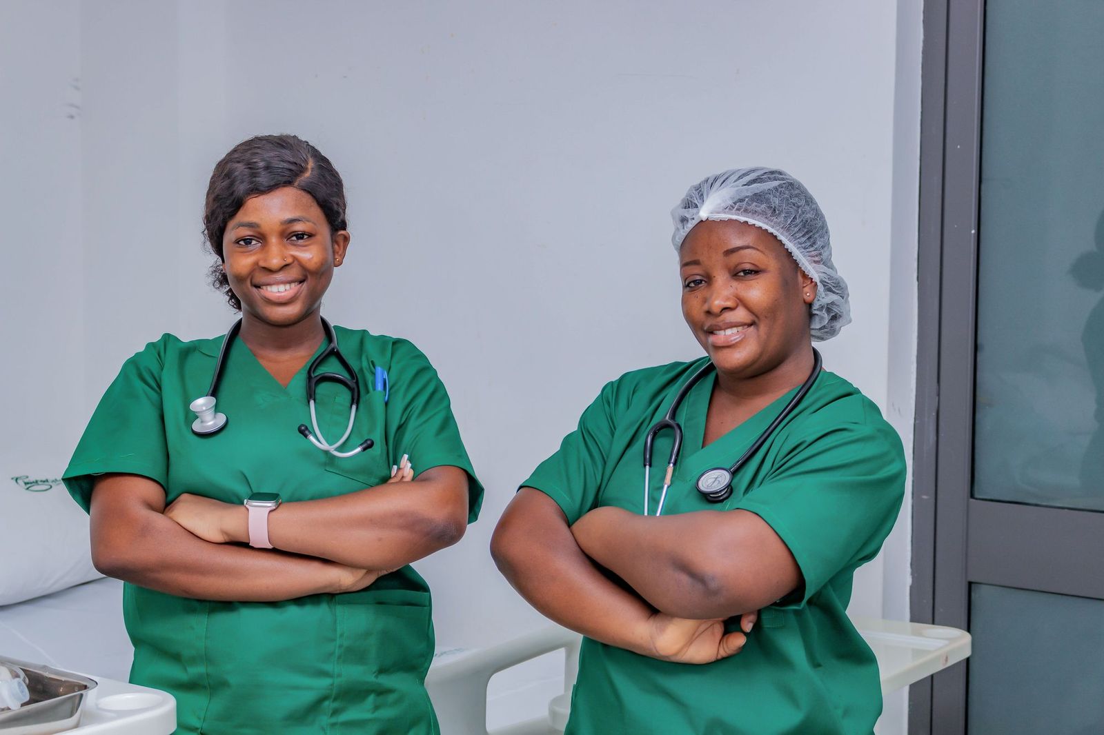 Two smiling nurses in green scrubs stand side by side with their arms crossed.