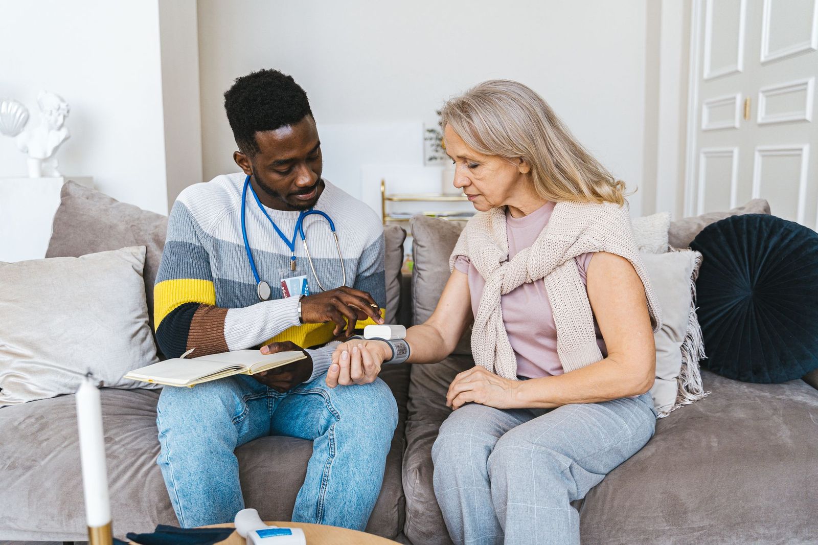 A smiling home health clinician checks an older woman’s blood pressure while they sit together on her living room sofa.